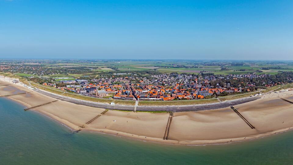 An aerial view of a coastal town with a beach, a fortified wall, and surrounding fields.