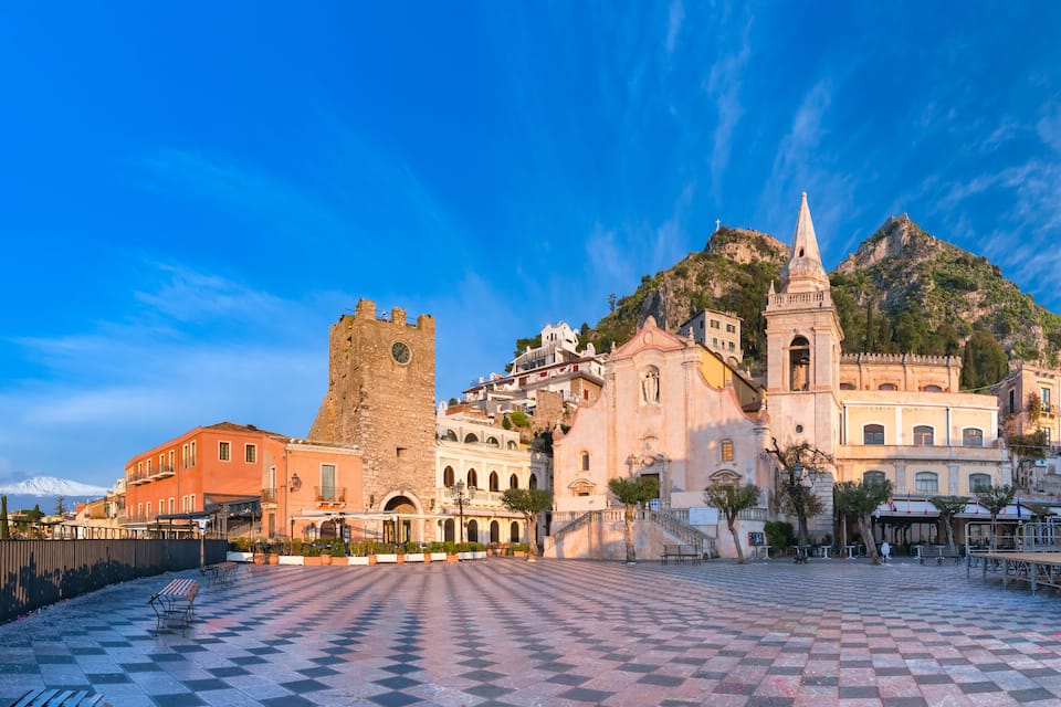 A historic square with a checkered pavement, a prominent tower, and a church with a bell tower, surrounded by Mediterranean architecture.