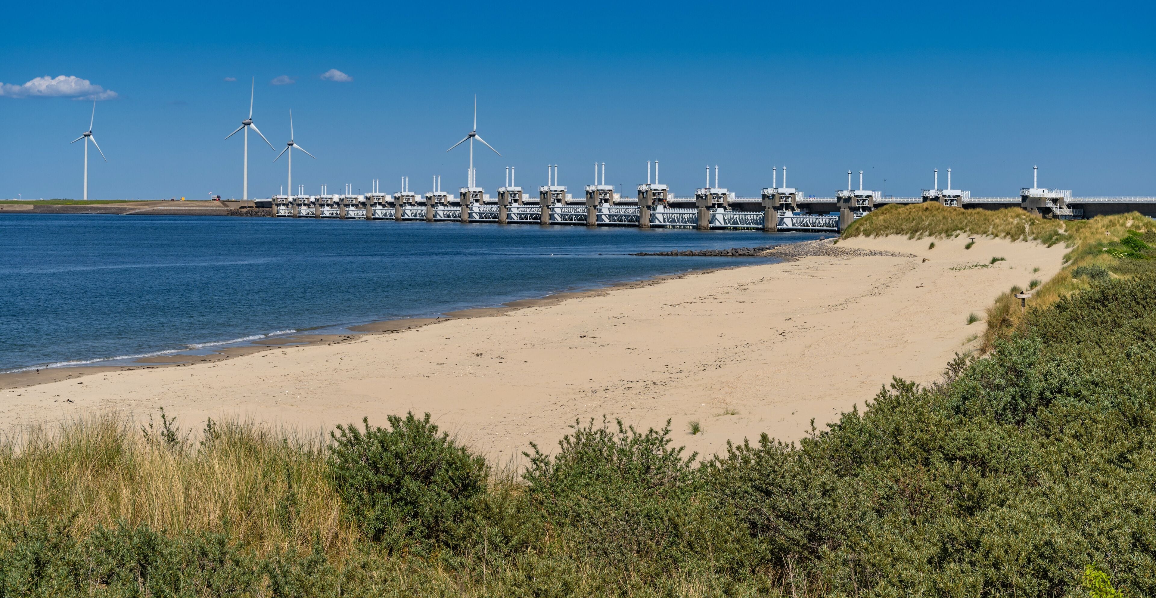 Un paisaje costero con una playa arenosa, turbinas eólicas y un puente sobre el agua.