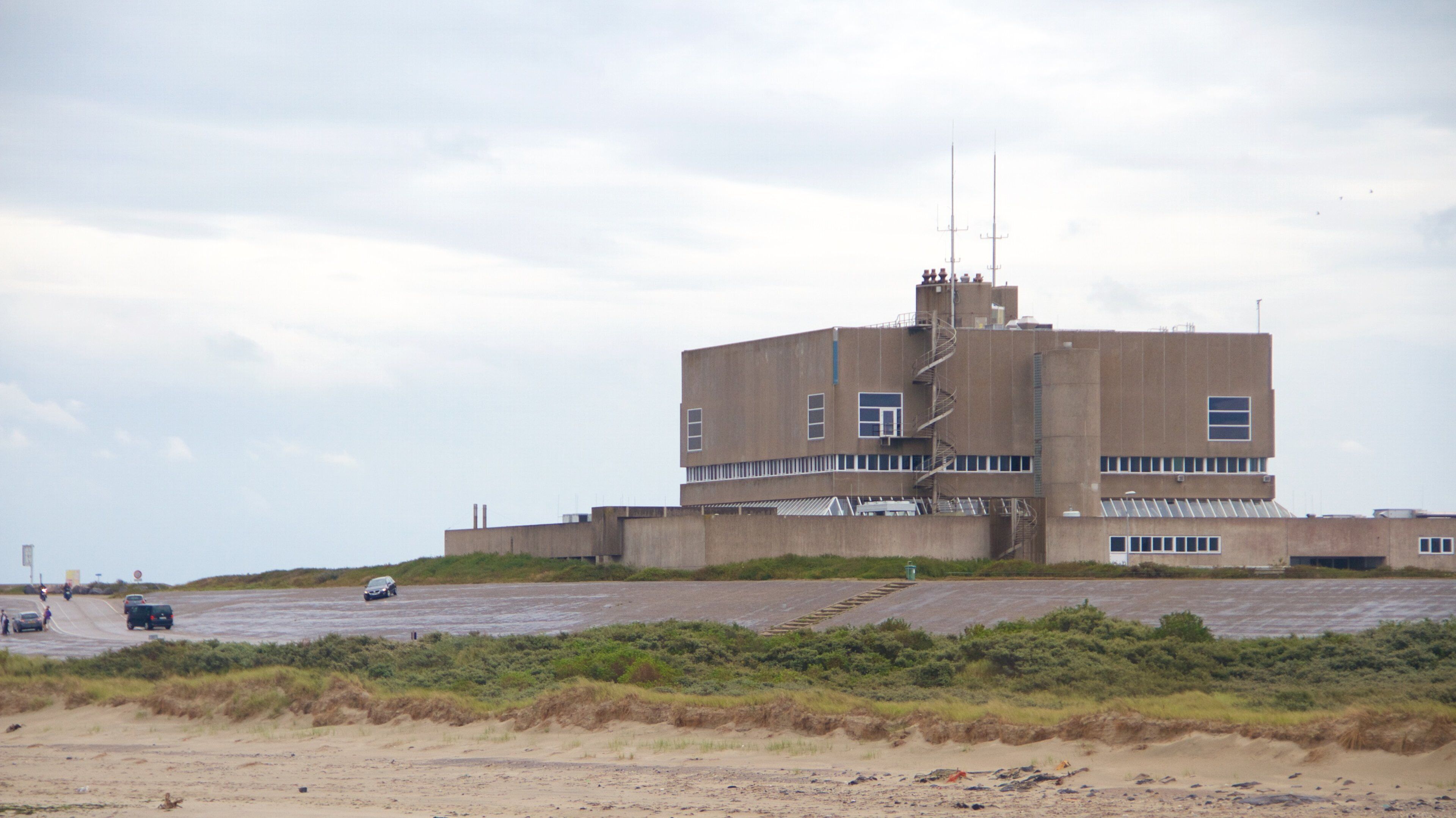 A large industrial building with a central tower and multiple windows, surrounded by a sandy area with sparse vegetation.