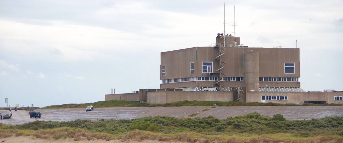 A large industrial building with a central tower and multiple windows, surrounded by a sandy area with sparse vegetation.