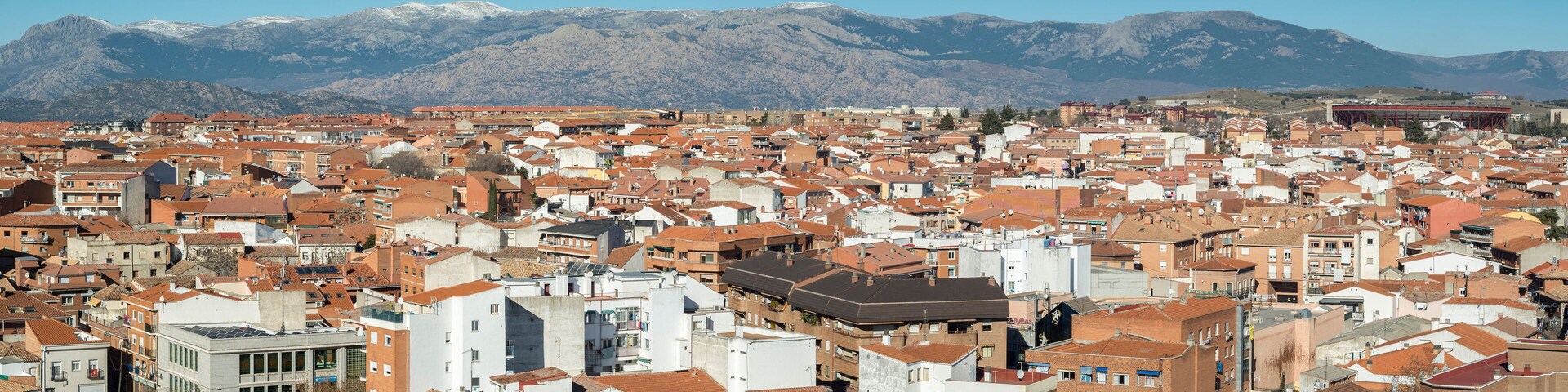 Un paisaje urbano con edificios de techos rojos y una cadena montañosa al fondo.
