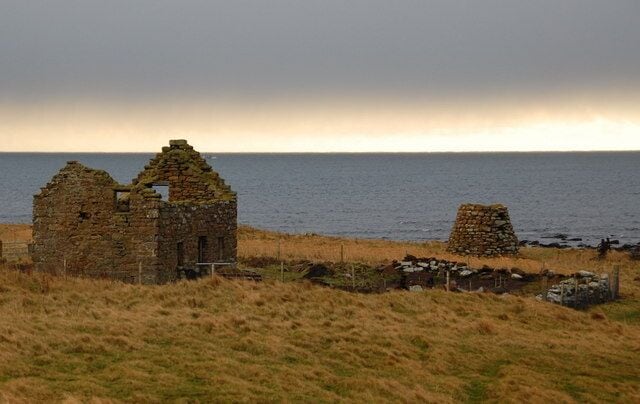 A coastal landscape with two stone ruins and a stone wall, surrounded by grassy terrain and the sea in the background.