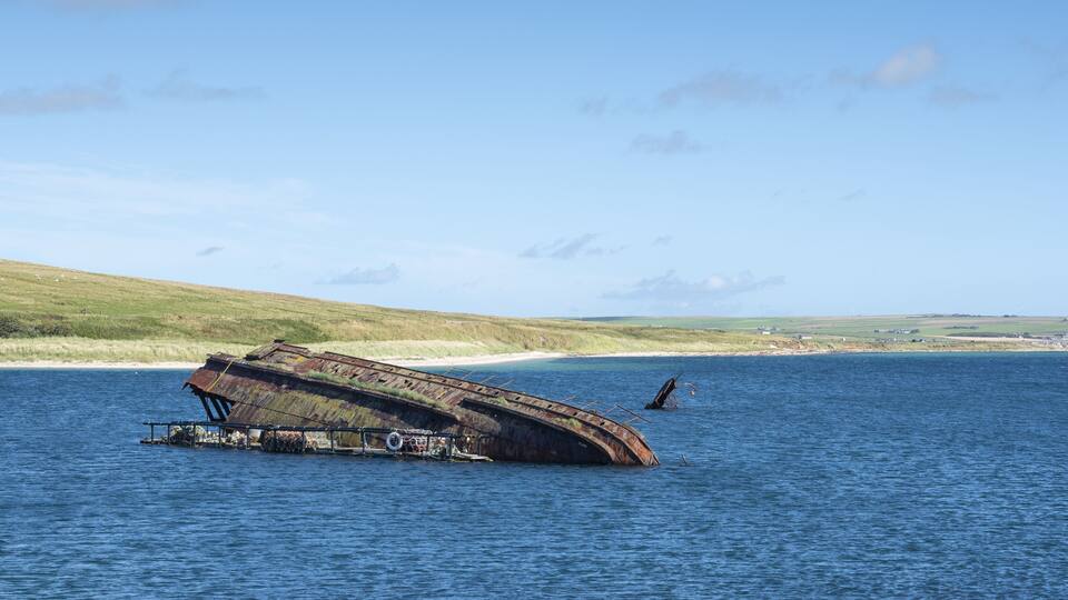 A sunken shipwreck in a body of water with a grassy shoreline and a clear sky in the background.