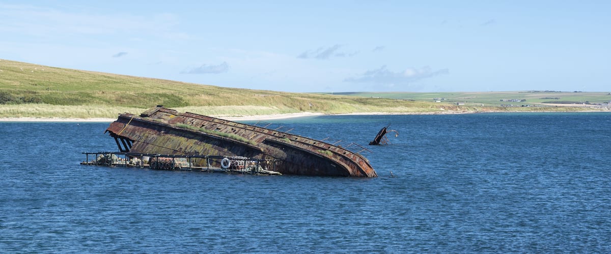 A sunken shipwreck in a body of water with a grassy shoreline and a clear sky in the background.