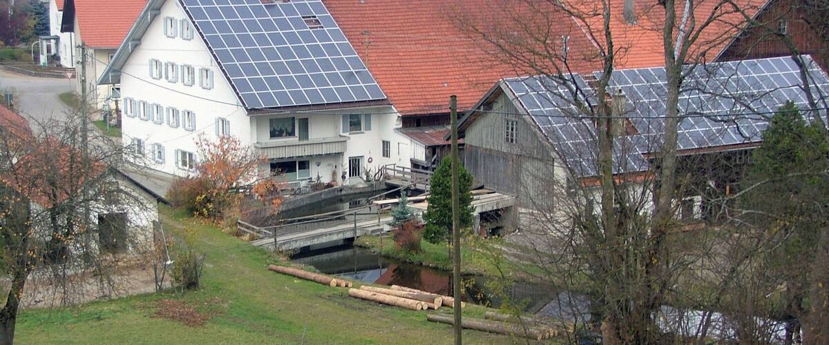 A rural landscape with a white house with solar panels, a wooden bridge, and a river.