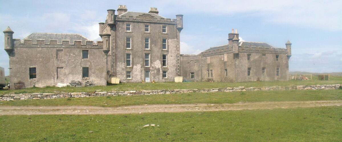 A large, historic stone building with multiple chimneys and windows, surrounded by a grassy field and a stone wall.