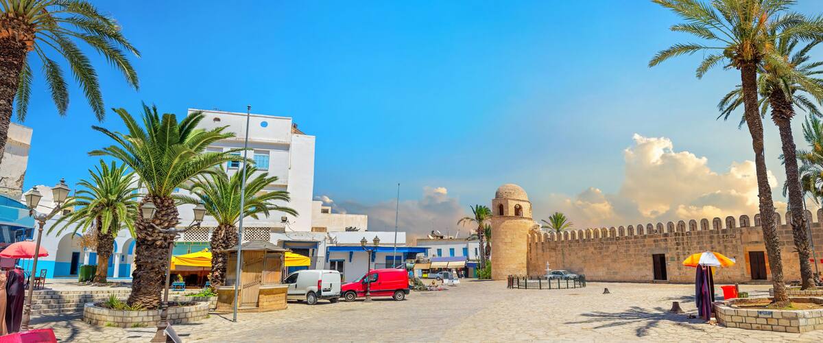 A historic square with palm trees, a white building with blue trim, and a stone wall with a tower.