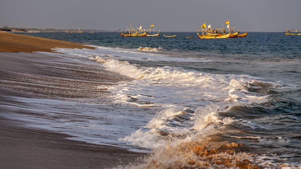 A beach with waves and several traditional boats in the background.