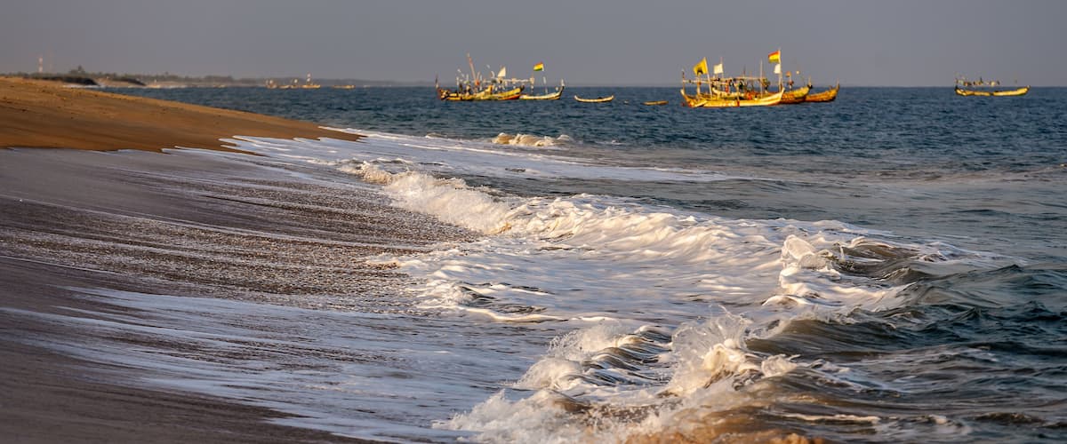 A beach with waves and several traditional boats in the background.