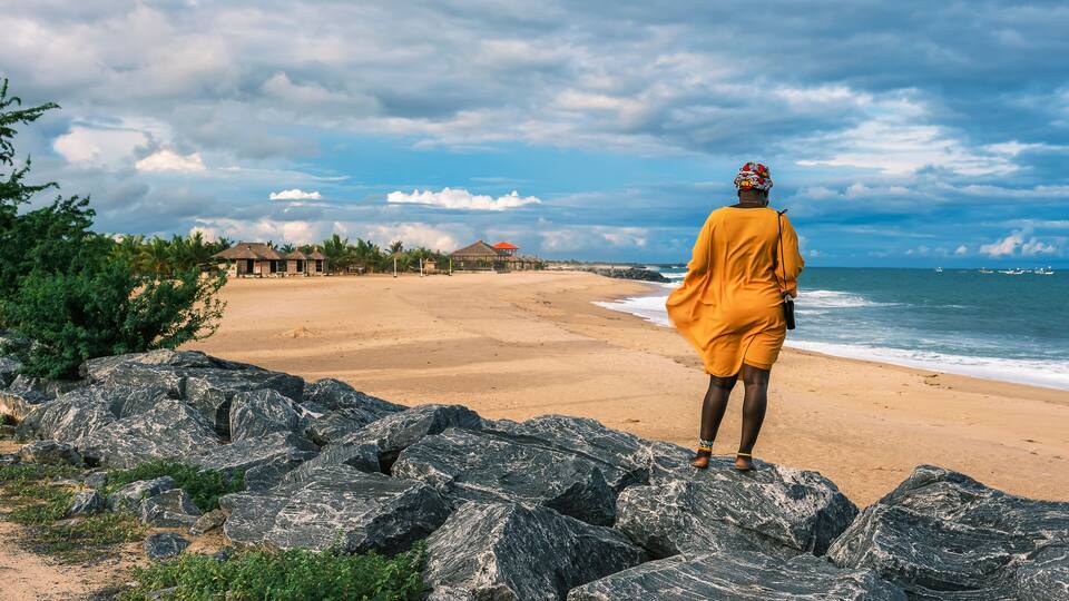 A beach with large rocks, a sandy shore, and a few buildings in the distance.