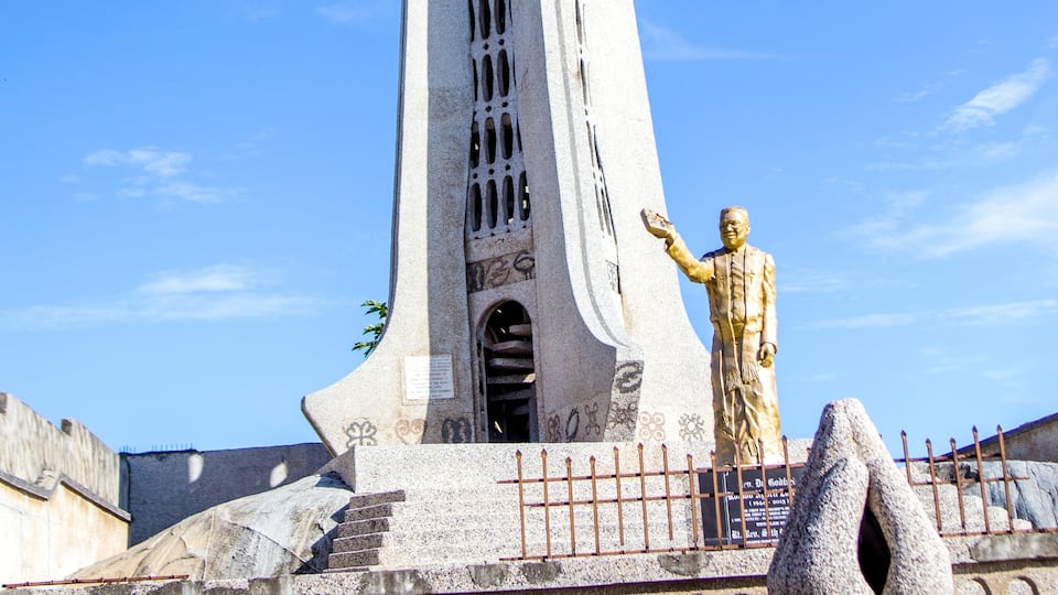 A tall white monument with a cross at the top, a statue of a man with outstretched arms, and a stone hand sculpture.