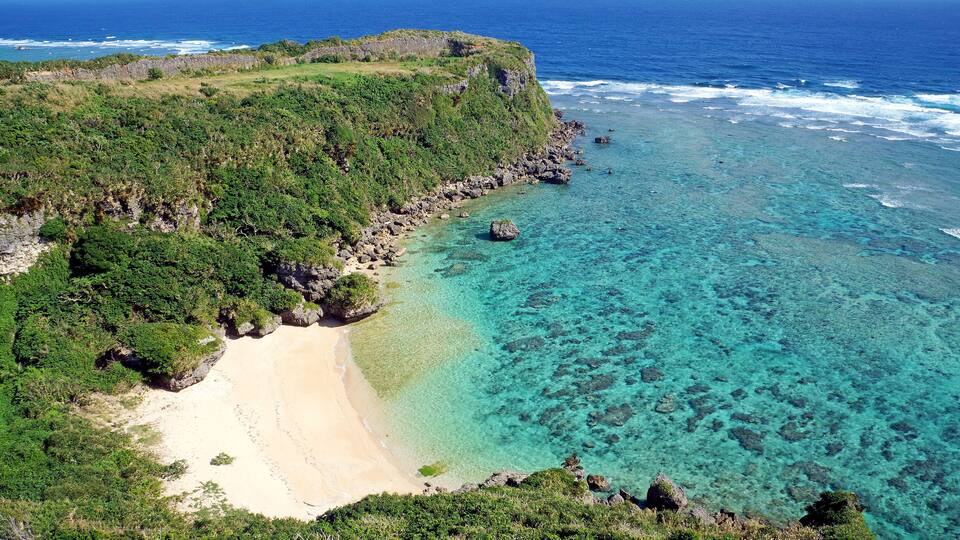 A coastal landscape with a sandy beach, clear turquoise waters, and lush green vegetation.