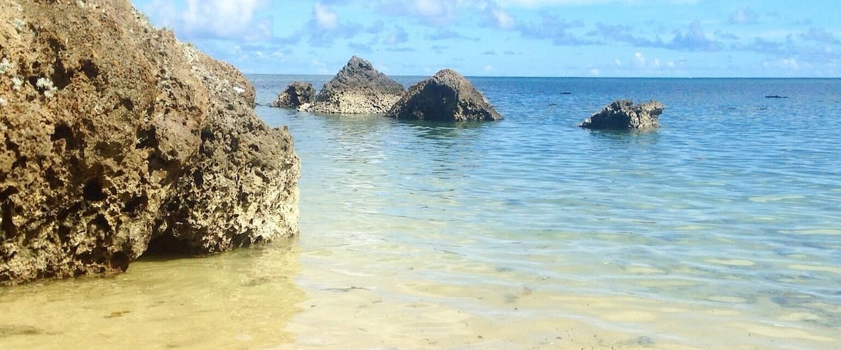 A coastal scene with clear shallow waters, rocky formations, and distant islands.