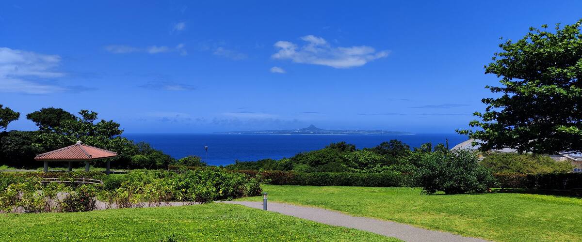 A park with a gazebo, a pathway, lush greenery, and a view of the ocean and distant land.