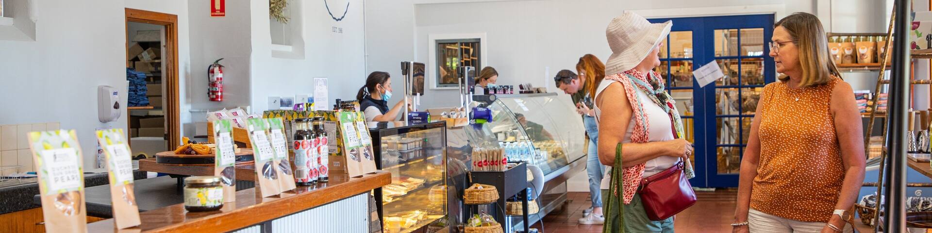 A cheese shop interior with a display counter, customers, and a sign reading "HANDMADE cheese from Victoria's HILLY COUNTRY".