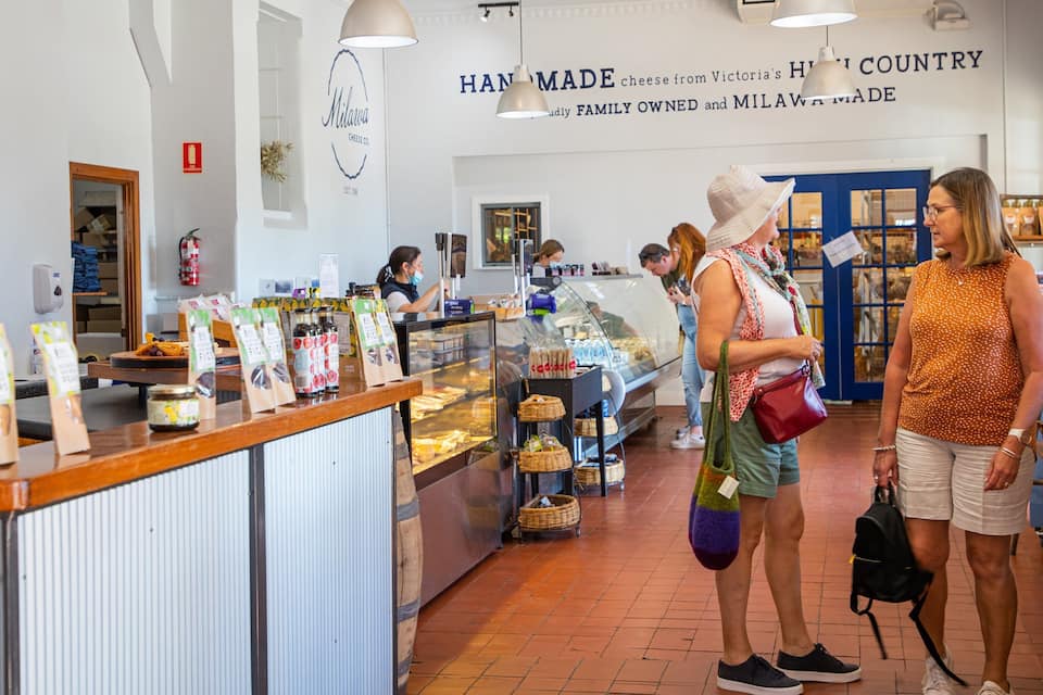 A cheese shop interior with a display counter, customers, and a sign reading "HANDMADE cheese from Victoria's HILLY COUNTRY".