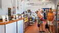 A cheese shop interior with a display counter, customers, and a sign reading "HANDMADE cheese from Victoria's HILLY COUNTRY".