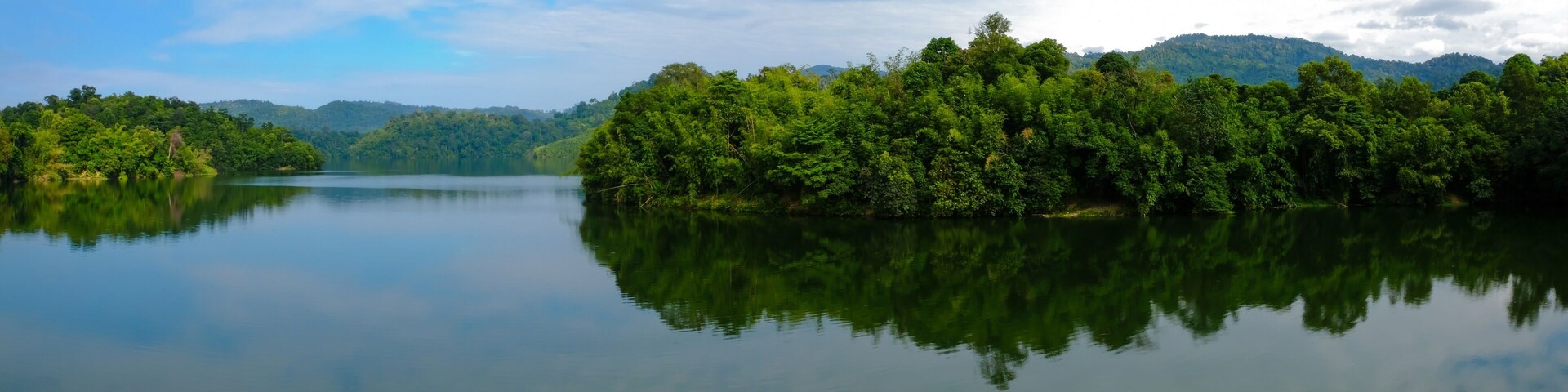 Un lago tranquilo rodeado de un bosque denso y montañas bajo un cielo parcialmente nublado.