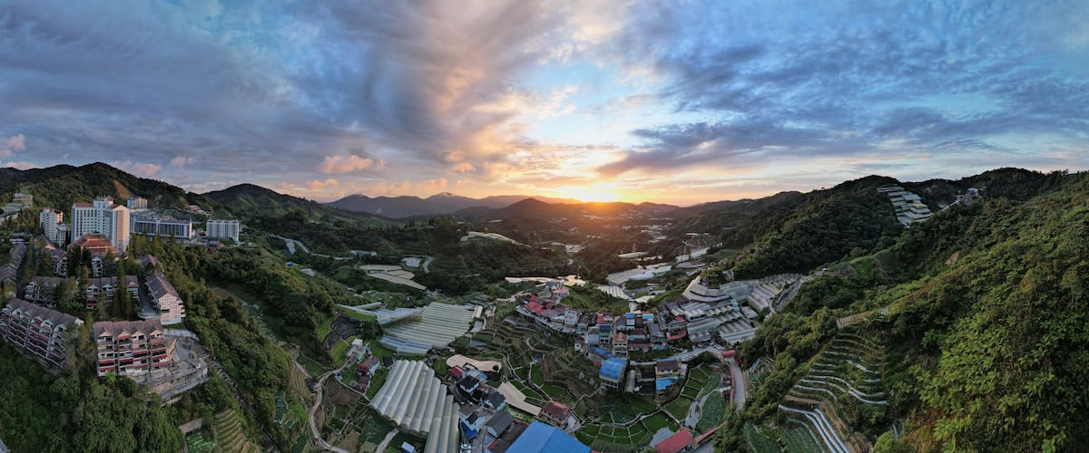 A panoramic view of a valley with terraced fields, residential buildings, and a sunset.