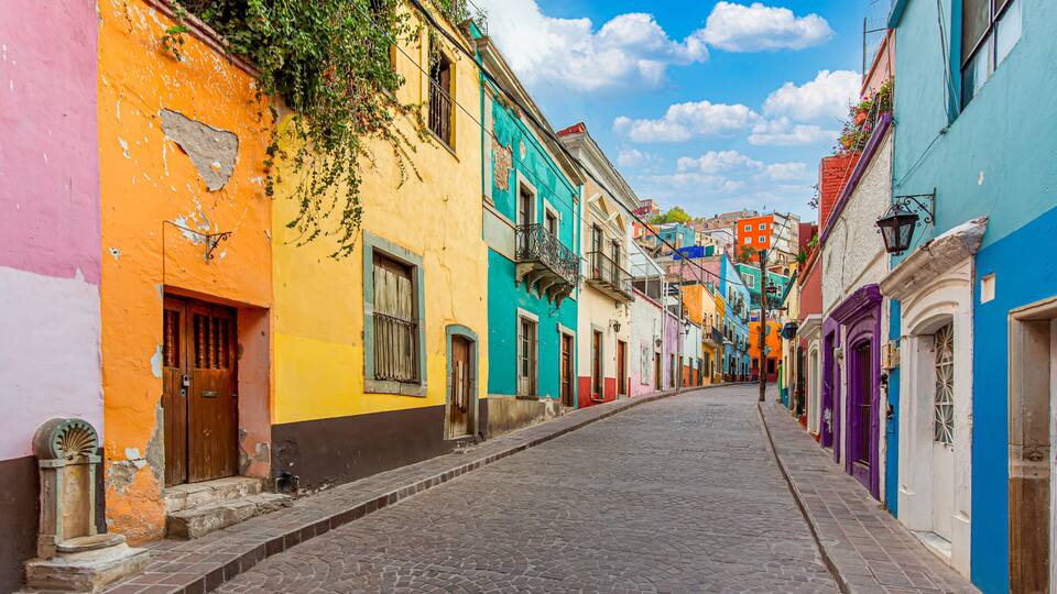 A cobblestone street lined with colorful buildings, a clear blue sky, and a few balconies with plants.