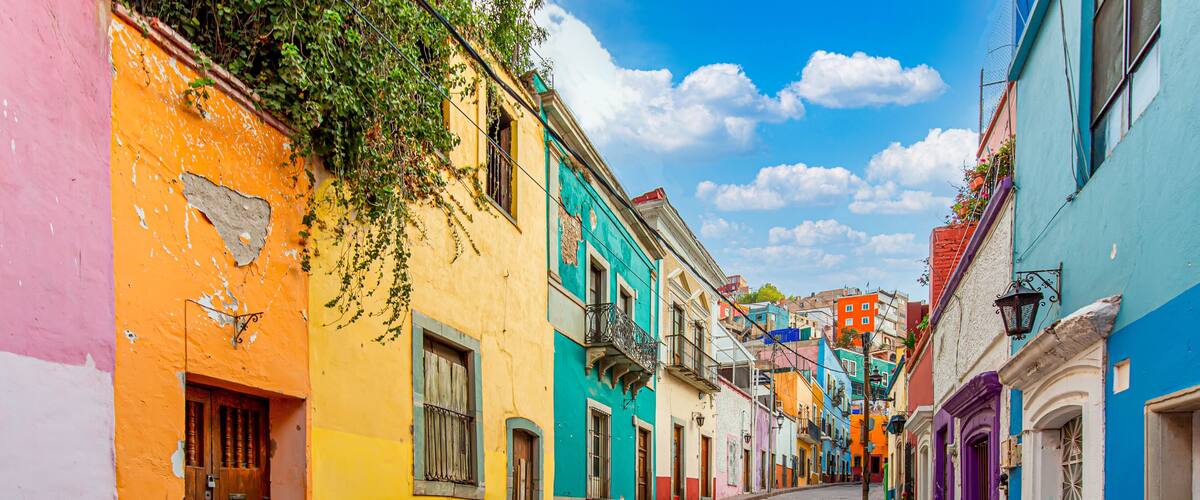 A cobblestone street lined with colorful buildings, a clear blue sky, and a few balconies with plants.