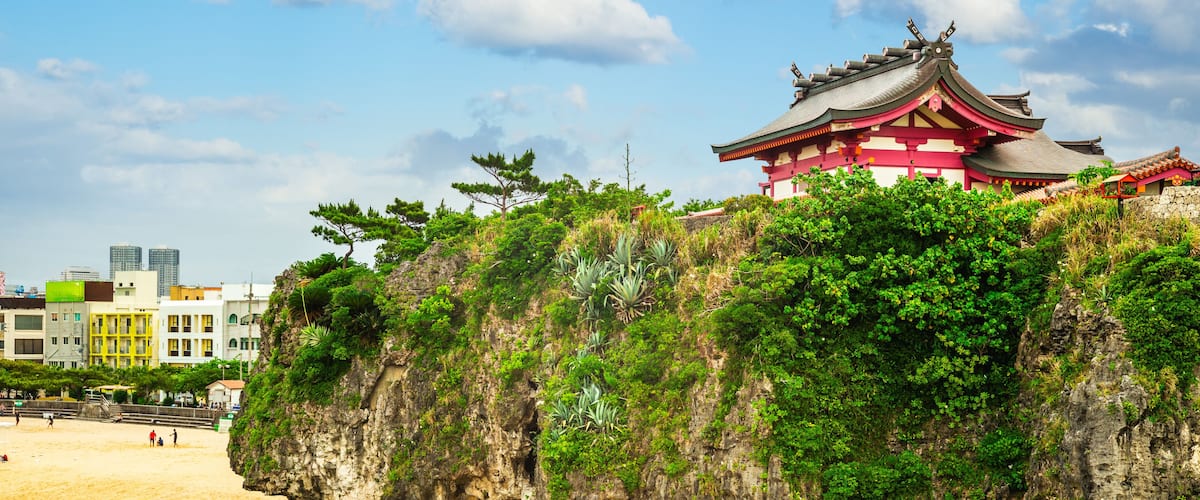 A traditional Japanese temple on a cliff overlooking a beach with a cityscape in the background.