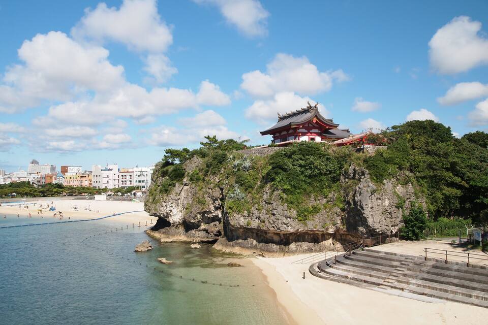 A traditional Japanese temple on a cliff overlooking a beach with clear blue water.