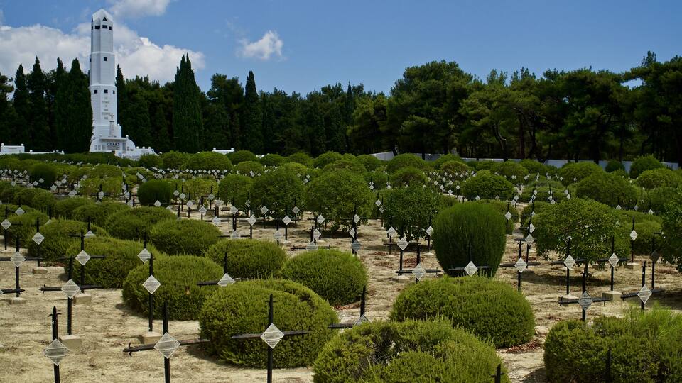 A cemetery with neatly trimmed bushes and gravestones, a white tower, and a forest in the background.
