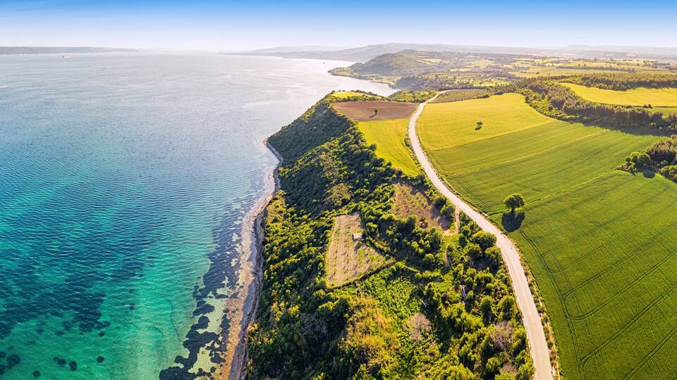 A coastal landscape with a clear view of the coastline, green fields, and a road leading to the sea.