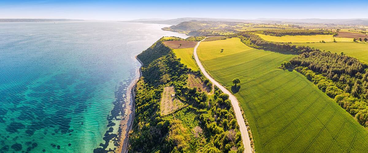 A coastal landscape with a clear view of the coastline, green fields, and a road leading to the sea.