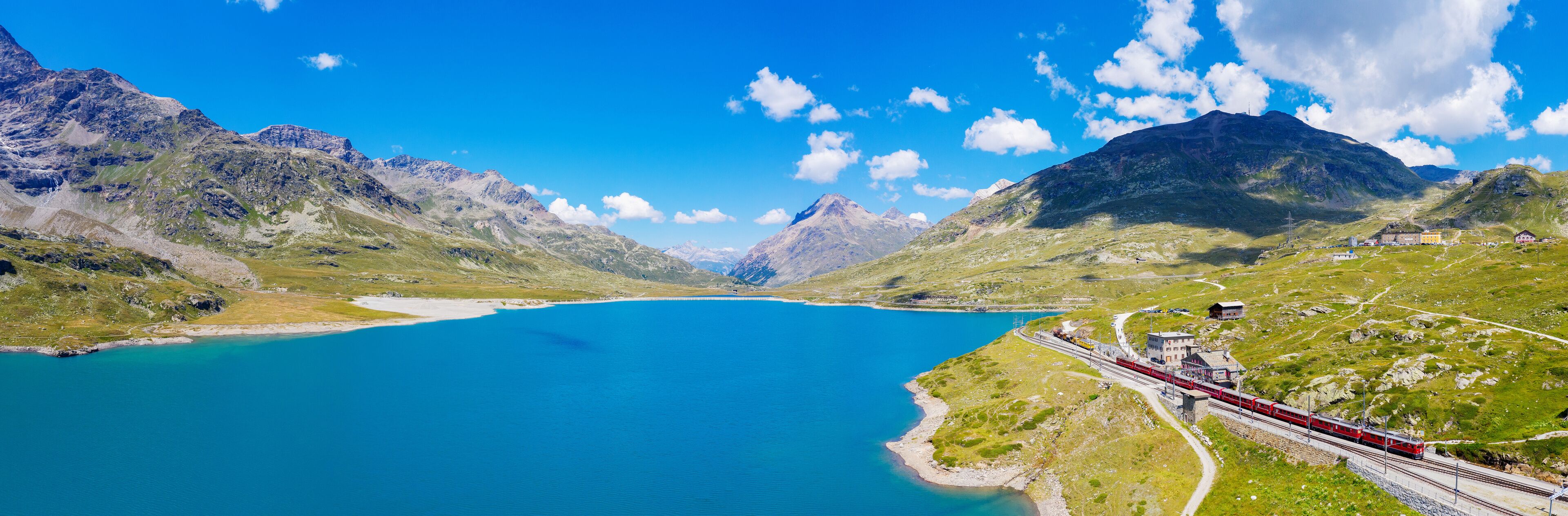 Un tren rojo viajando sobre rieles junto a un lago turquesa rodeado de montañas.