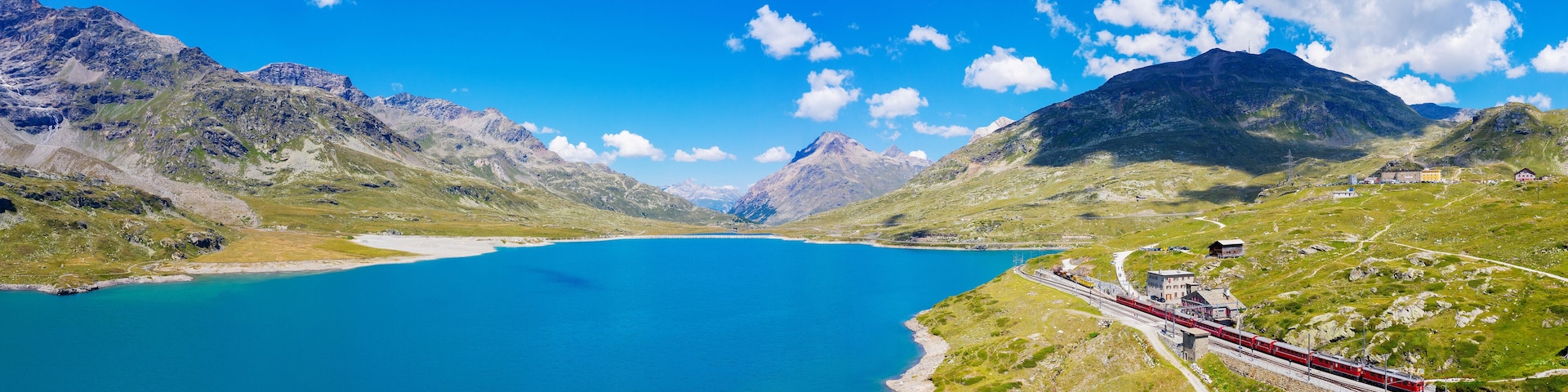 Un tren rojo viajando sobre rieles junto a un lago turquesa rodeado de montañas.