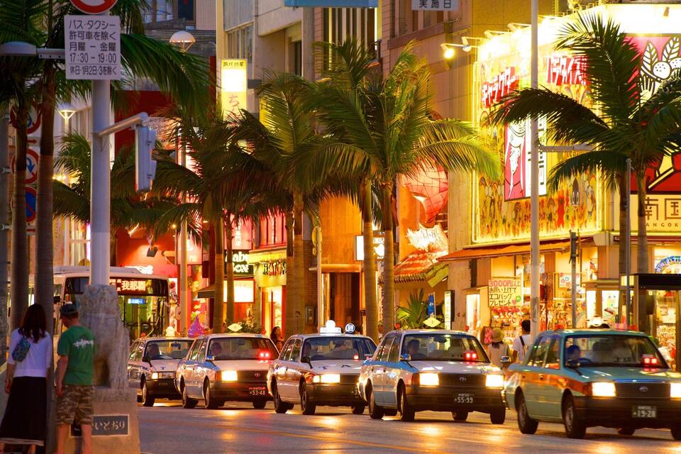 A busy urban street lined with palm trees, taxis, and brightly lit shops.