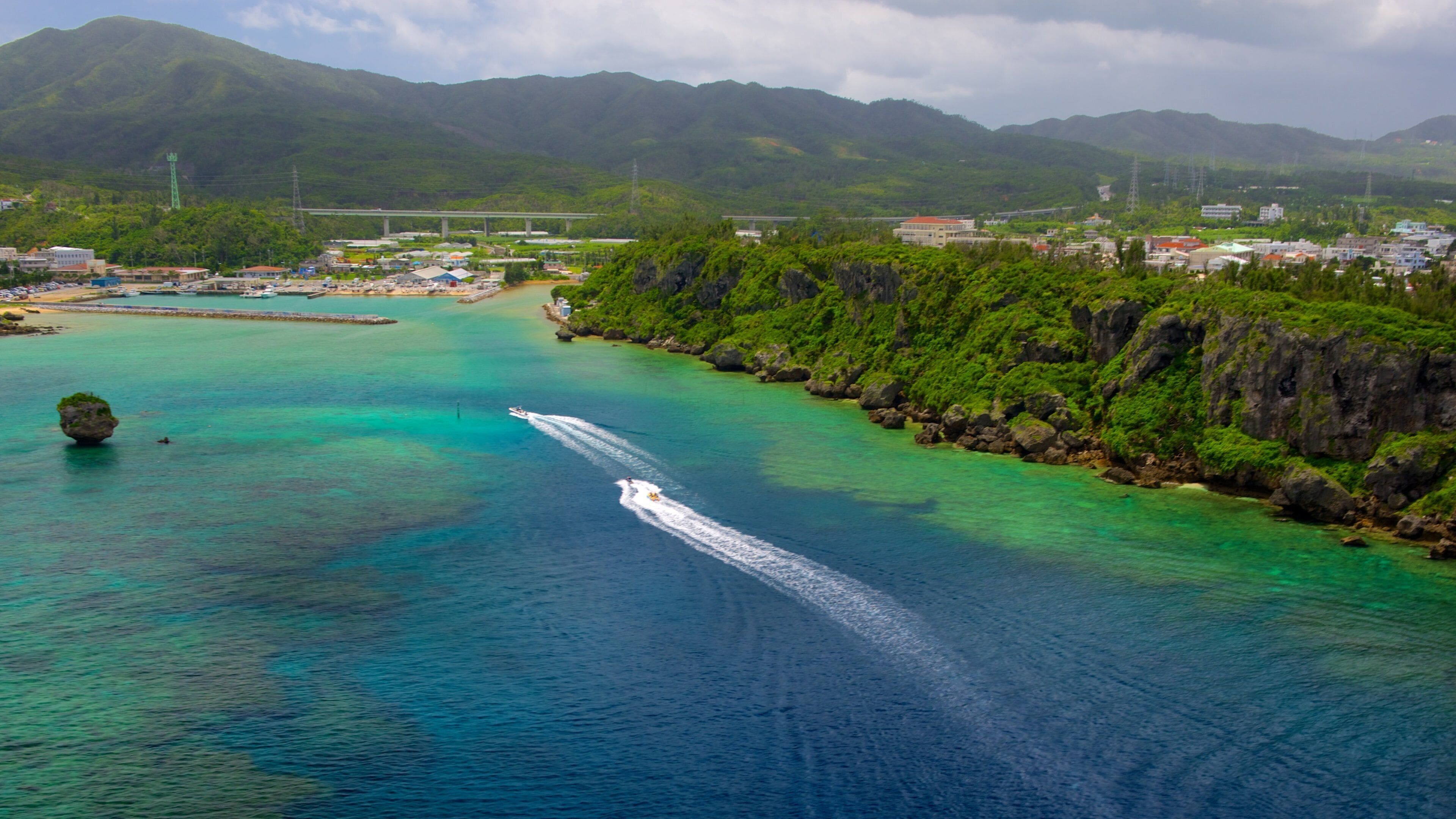 A coastal area with clear turquoise waters, a boat creating a wake, and lush green hills in the background.