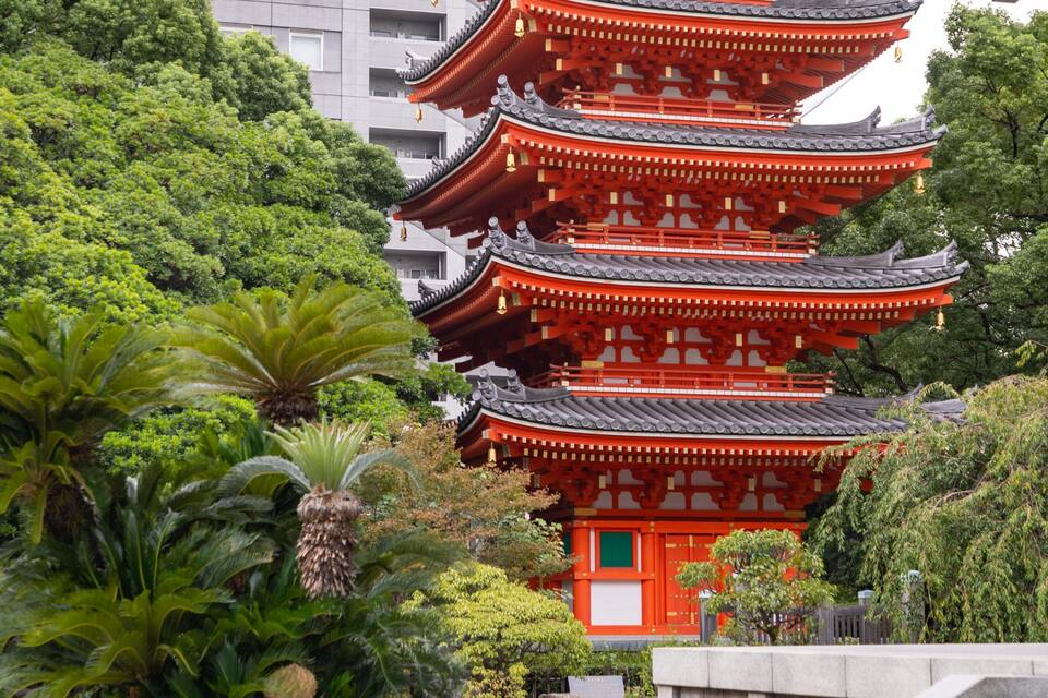 A traditional Japanese pagoda with multiple tiers, surrounded by lush greenery and modern buildings in the background.