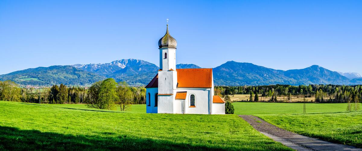 A white church with a red roof and a tall tower, surrounded by green fields and mountains in the background.
