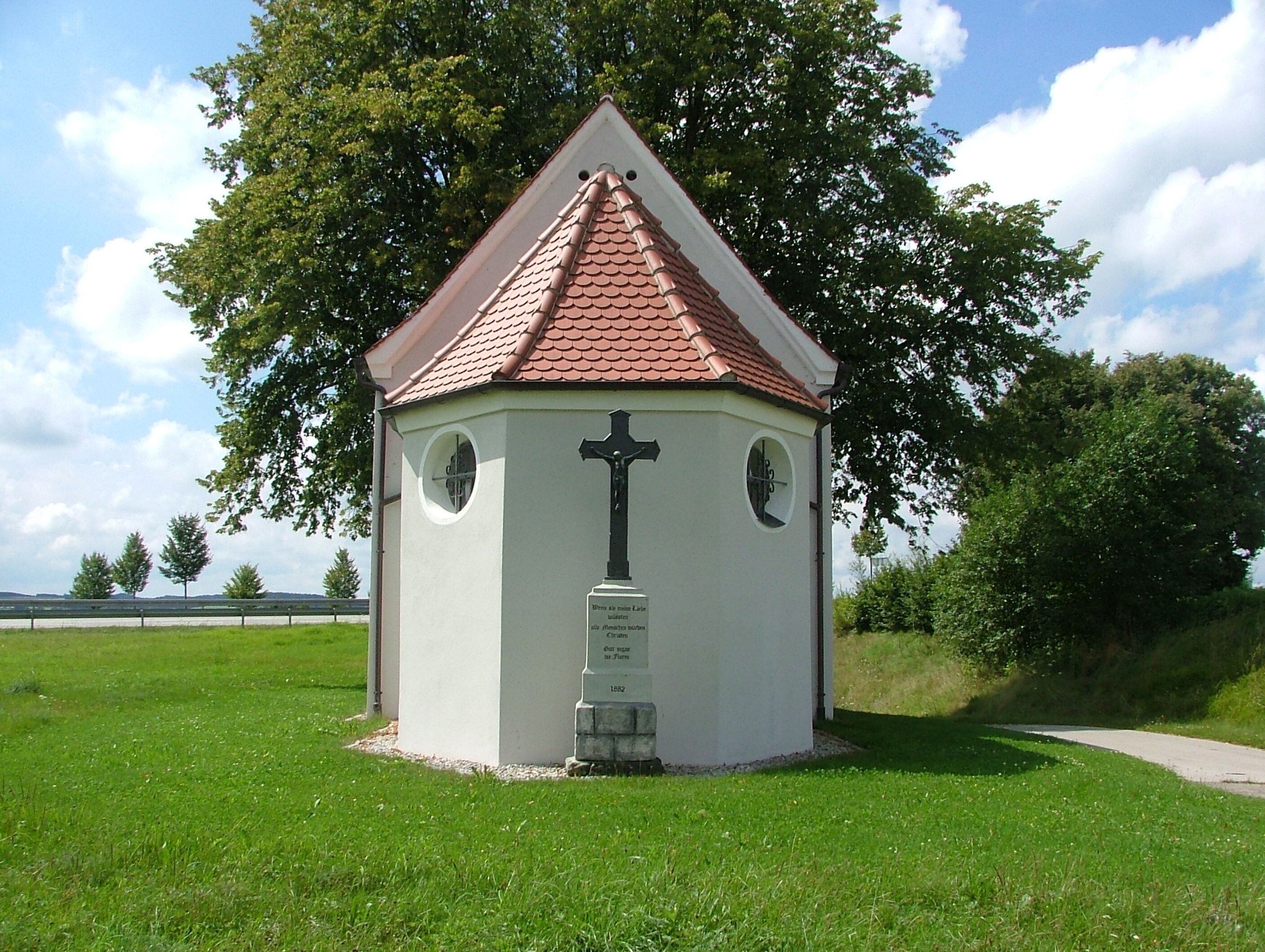 A small white chapel with a red roof and a cross, surrounded by green grass and trees.