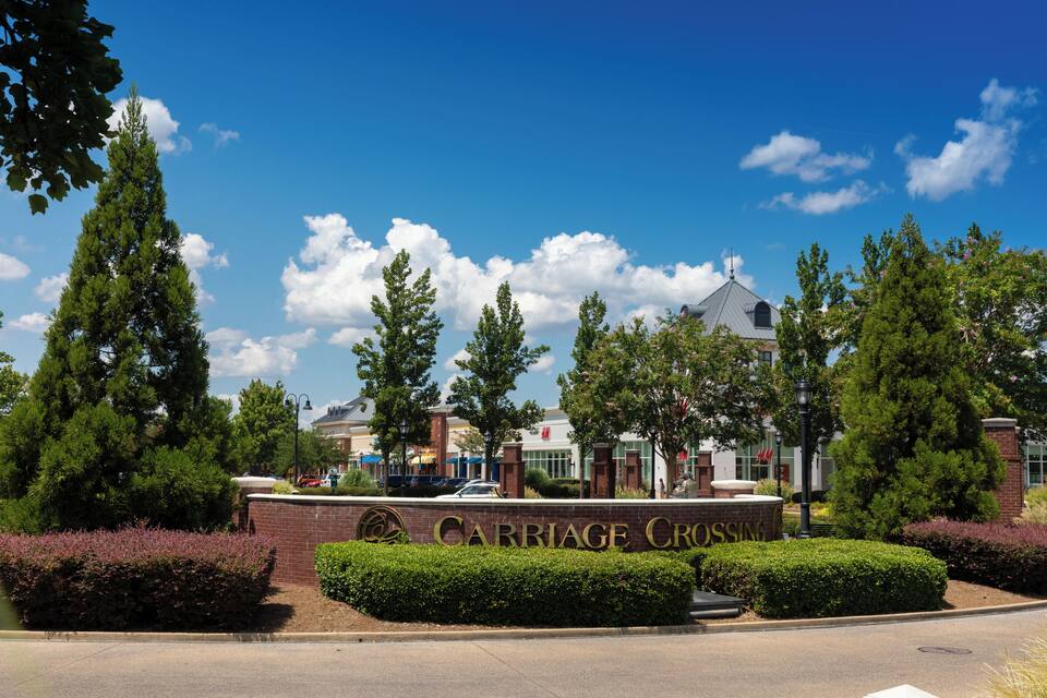 A brick entrance with "Carriage Cross" signage, surrounded by well-maintained hedges and tall trees.