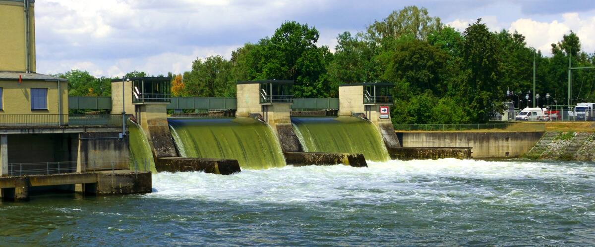 A dam with water flowing over it, surrounded by greenery and buildings.