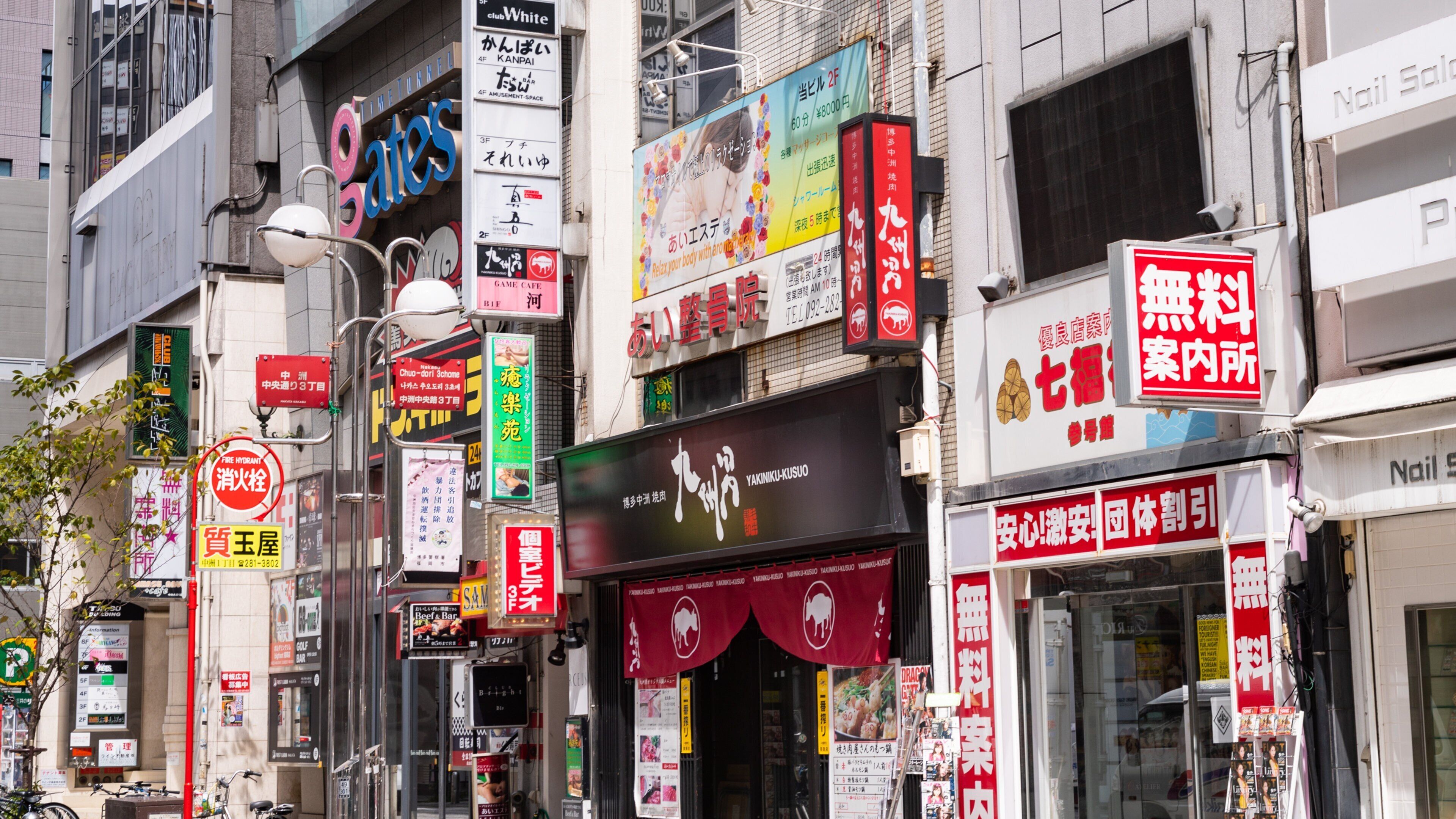 Eine belebte Stadtstraße mit verschiedenen Geschäften, darunter ein Café für Spiele, ein Nagelstudio und ein Restaurant.