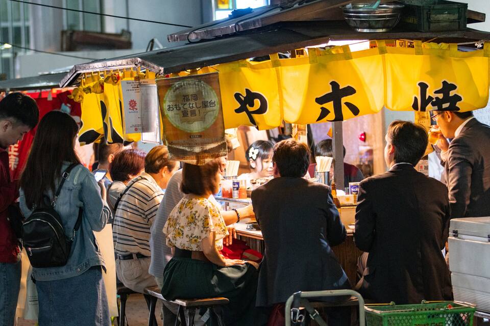 Un puesto de comida callejera concurrido, con señalización amarilla y una multitud de gente.