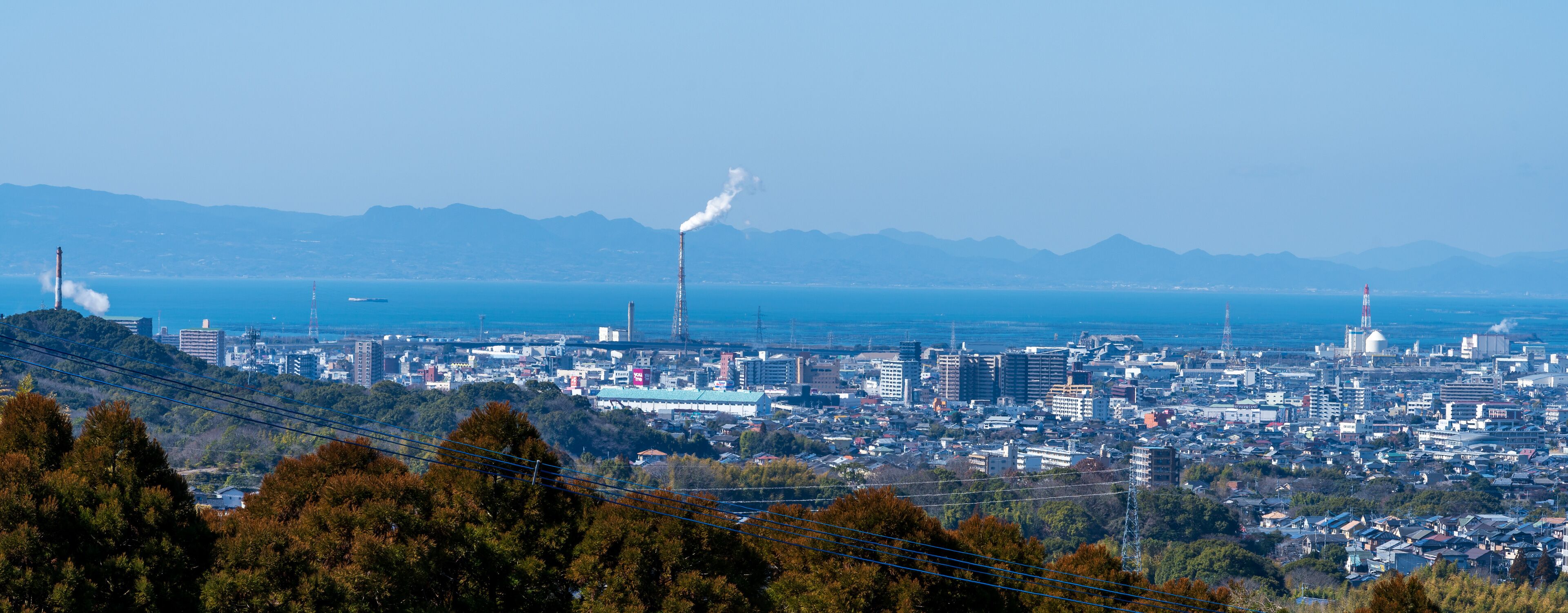 A coastal cityscape with industrial buildings, a prominent smokestack, and mountains in the background.