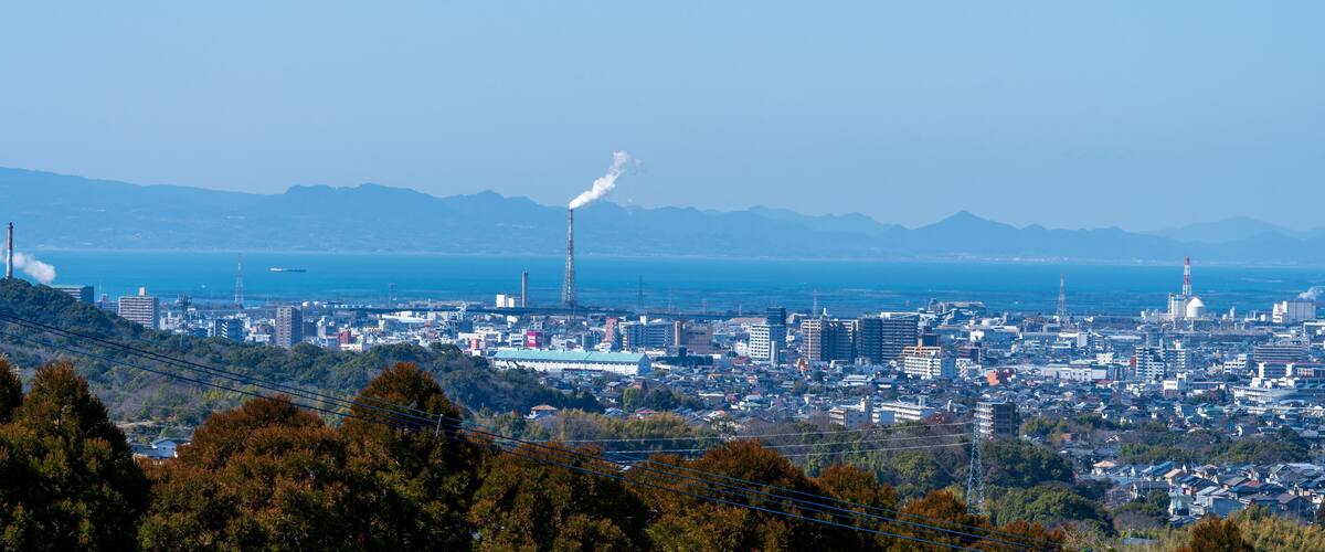 A coastal cityscape with industrial buildings, a prominent smokestack, and mountains in the background.