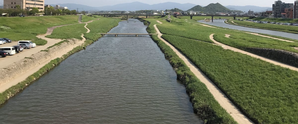 A river flowing through a city with green banks, a bridge, and distant mountains.