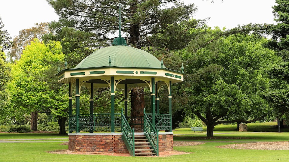 A gazebo with a green roof and brick base, surrounded by lush green trees and grass in a park.