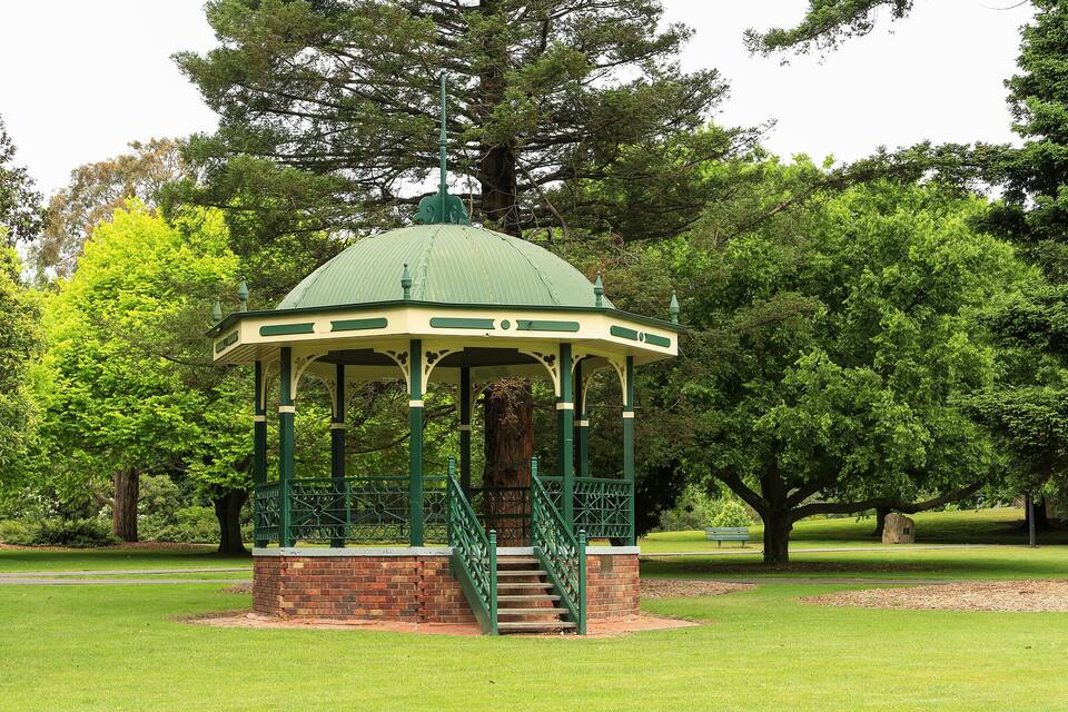 A gazebo with a green roof and brick base, surrounded by lush green trees and grass in a park.