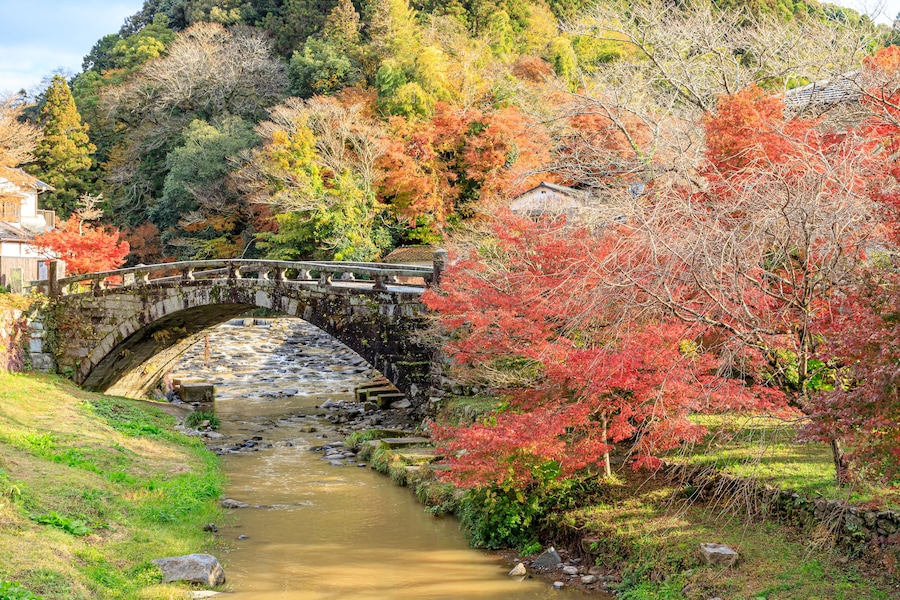 A stone bridge over a river, surrounded by autumn foliage and a hillside.