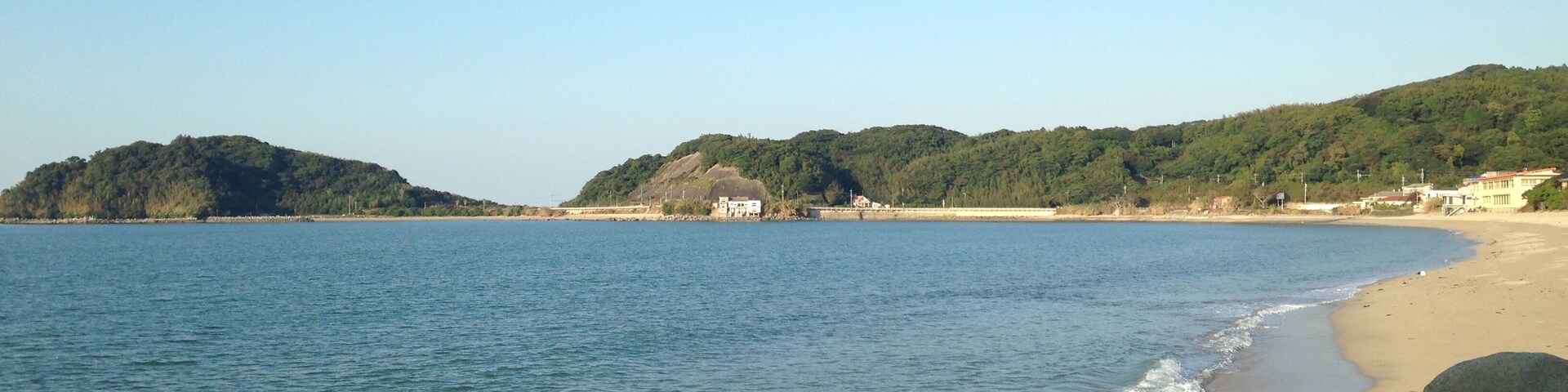 Uma costa rochosa com mar calmo, uma praia de areia e colinas ao fundo.