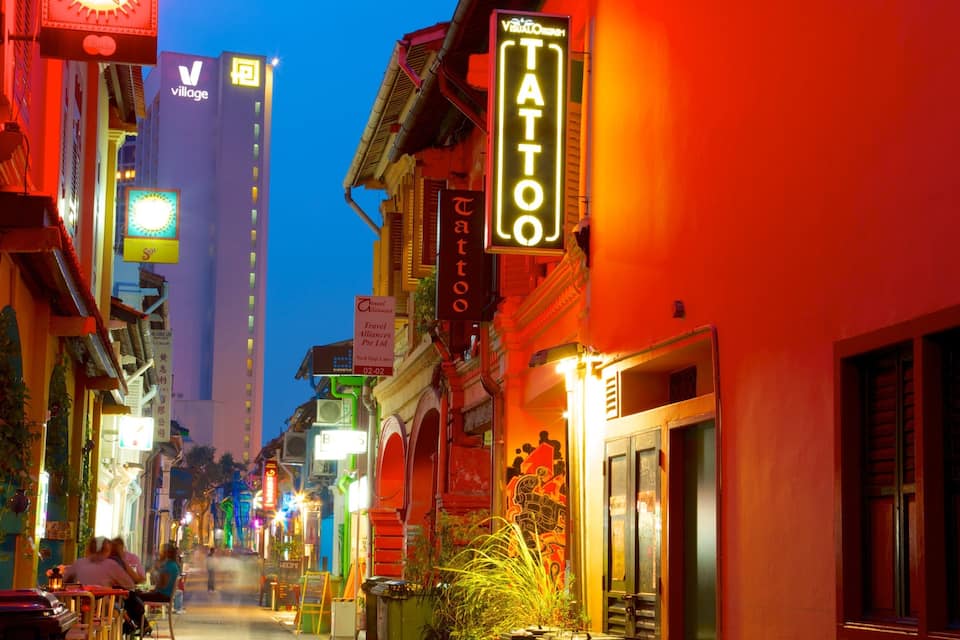 A brightly lit street with a tattoo parlor and a tall building with a 'village' sign.